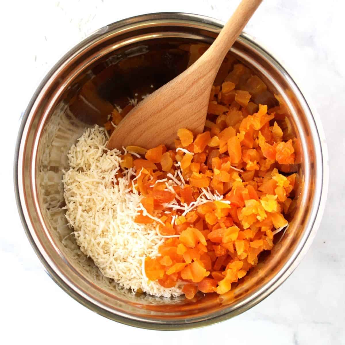 chopped apricots and shredded coconut side by side in a stainless steel mixing bowl with a wooden spoon.