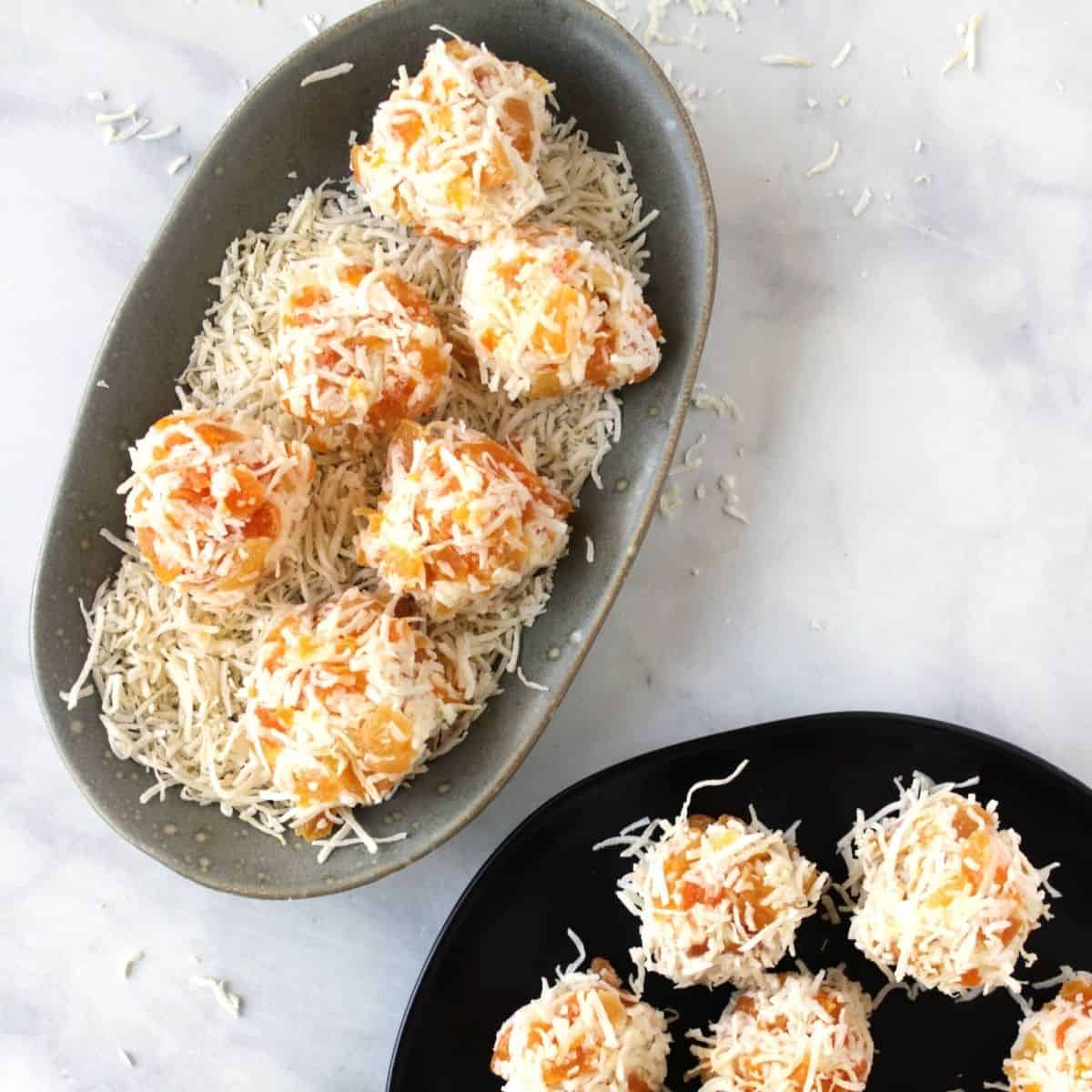 green oblong plate with six apricot balls sitting in a bed of shredded coconut with a black plate with more apricot balls showing partially in the corner.