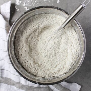 looking down into a large stainless steel bowl of flour with a whisk in it.