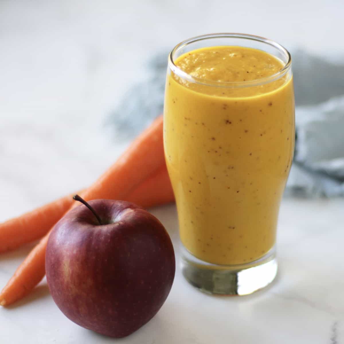 deep red apple and a couple carrots sit by a glass of golden apple carrot smoothie.