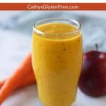 Glass of golden smoothie by partially shown apple and carrots.