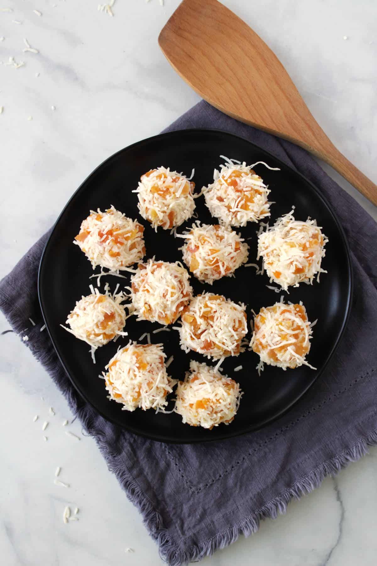 looking down onto a black plate with several coconut-coated apricot balls sitting on a lavender napkin with a wooden spatula to the side.