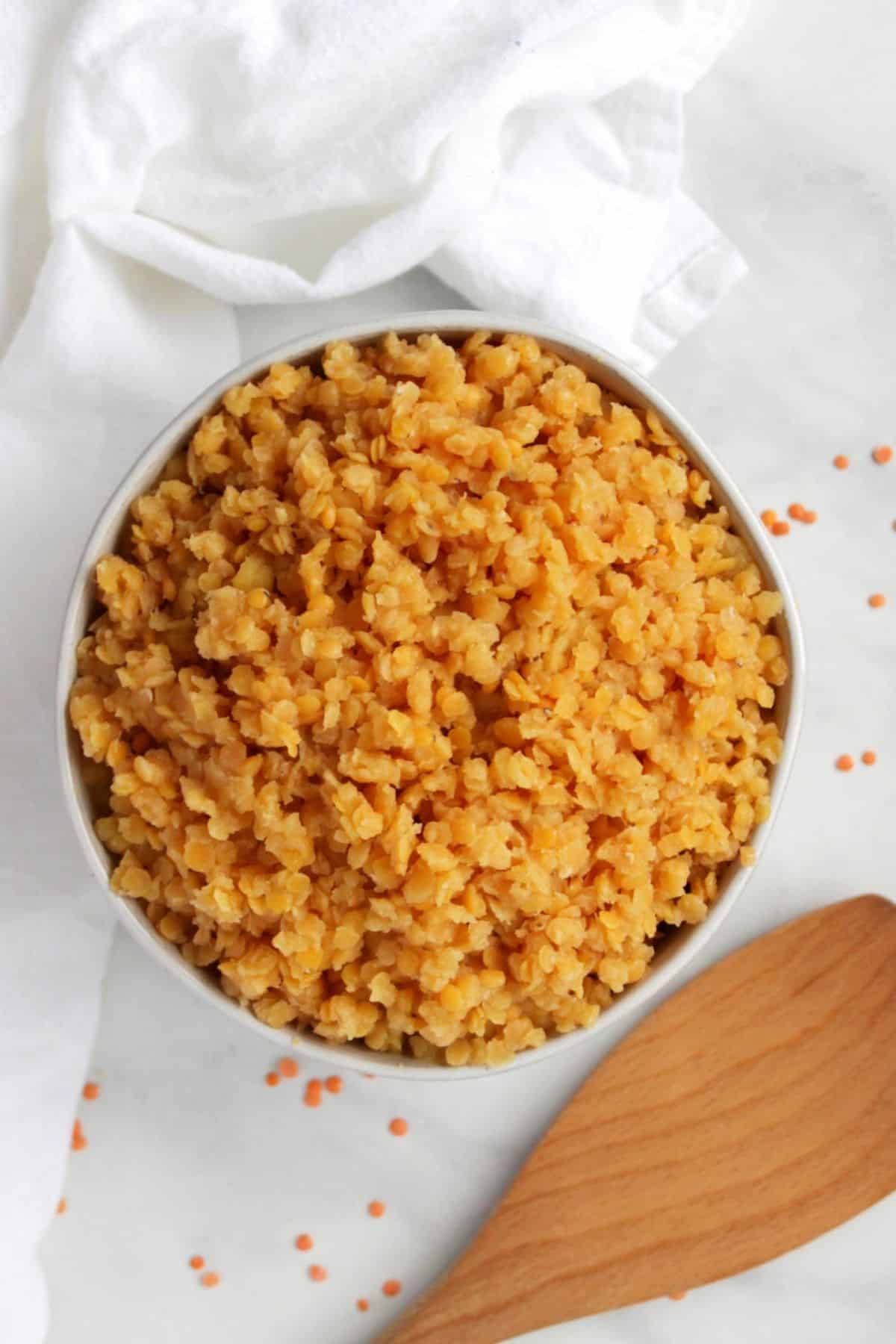 looking down into a heaping bowl of cooked red lentils with white cloth on marble surface.