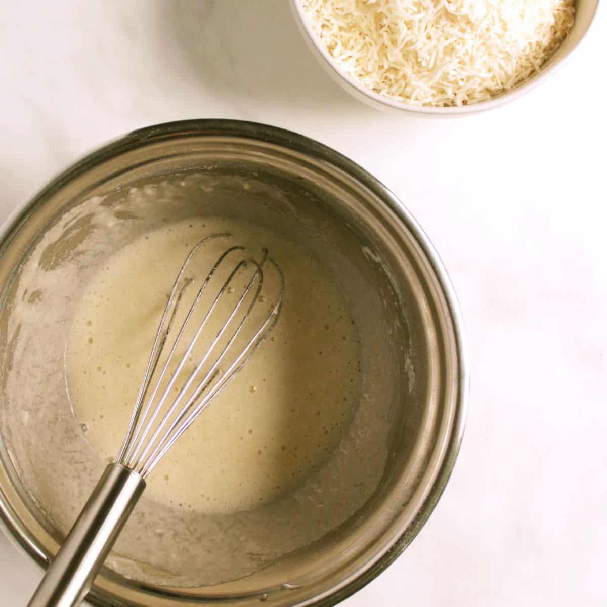 looking down into stainless steel mixing bowl with a whisk in some creamy batter, and a bowl of flaked coconut partially seen off the edge.