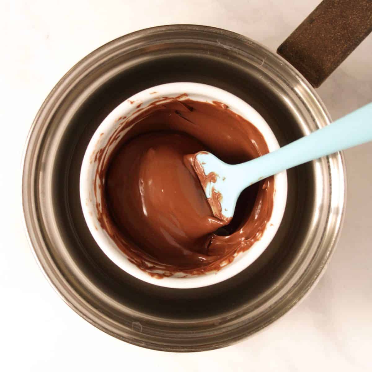 spatula stirs liquid chocolate in white bowl set in saucepan of water.