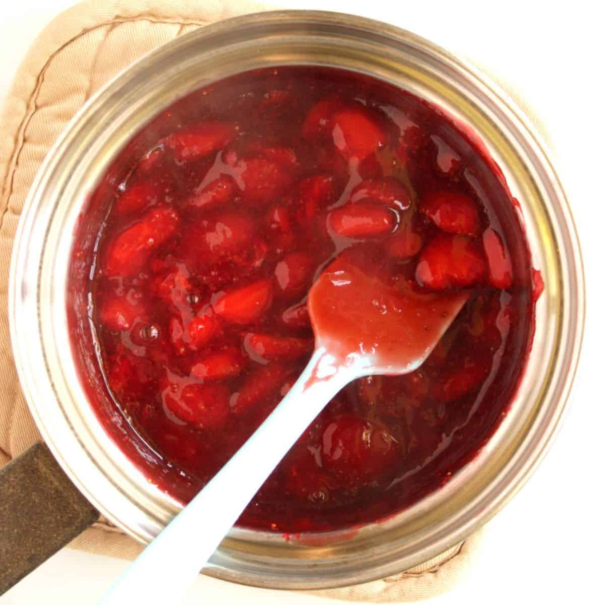 cooked strawberry chunks in red gel with pale blue spatula in small stainless saucepan.