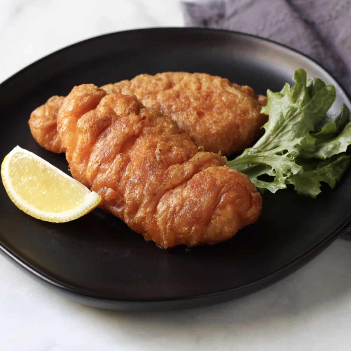 two pieces of crispy, golden-browned battered fish on a black plate with a wedge of lemon and a lettuce leaf.