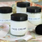 clear jars of white face cream with black lids sitting on cloth with pink ribbon.