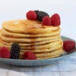 stack of pancakes on blue plate drizzled with syrup and garnished with berries.