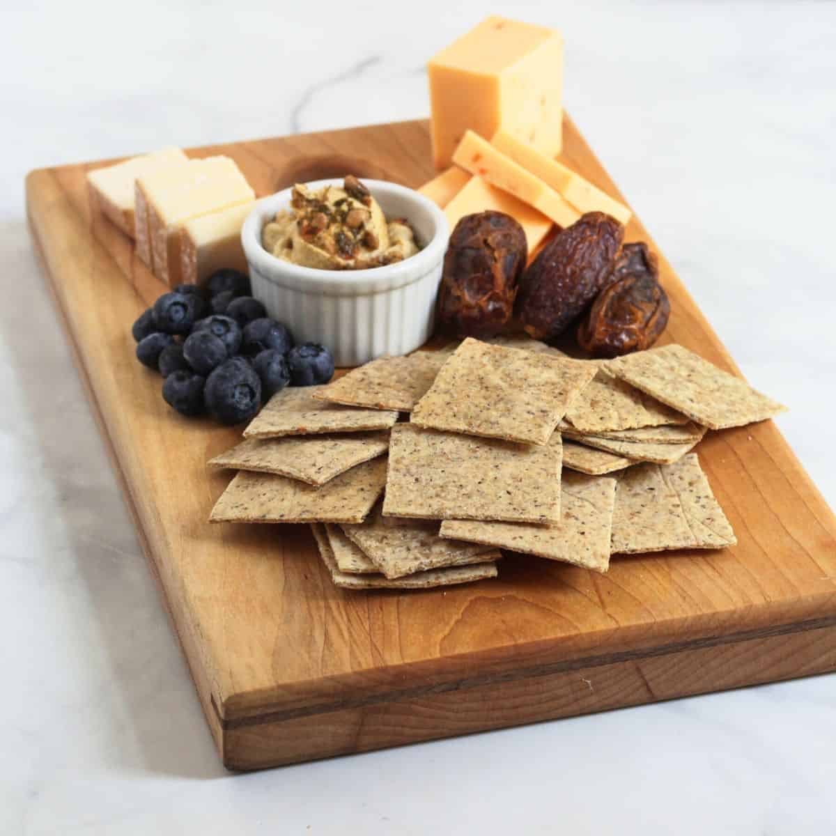 square crackers on wooden board with nuts, cheese, and fruit.