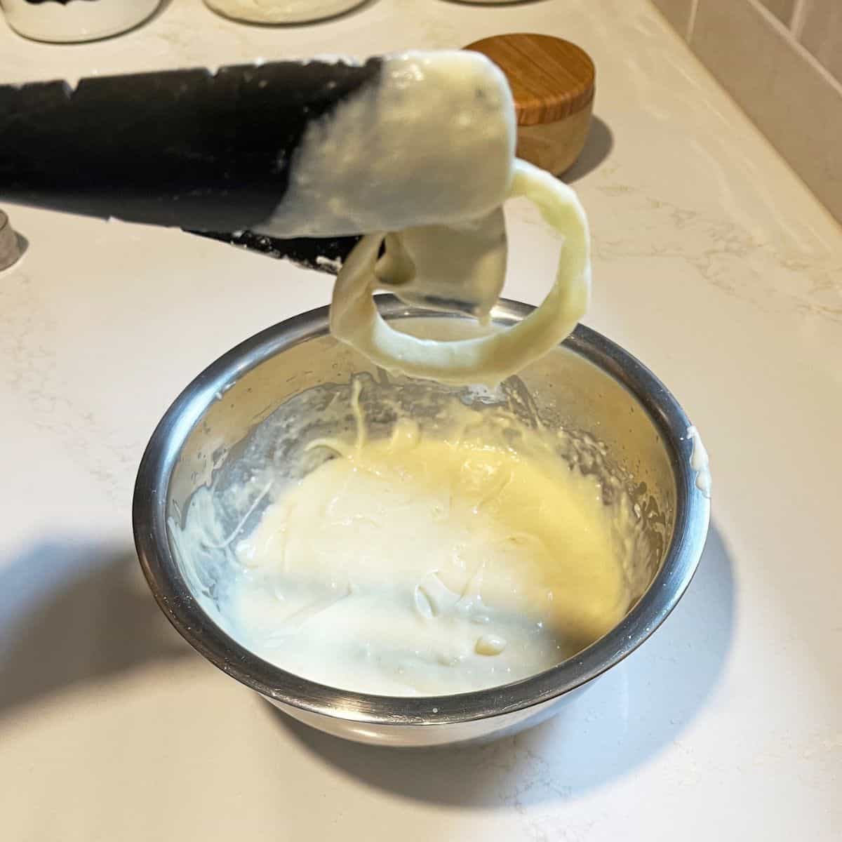 Batter-dipped onion ring is lifted from stainless steel bowl of batter.