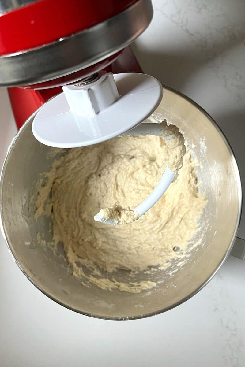 dough hook raised above soft sticky dough in bowl of stand mixer.