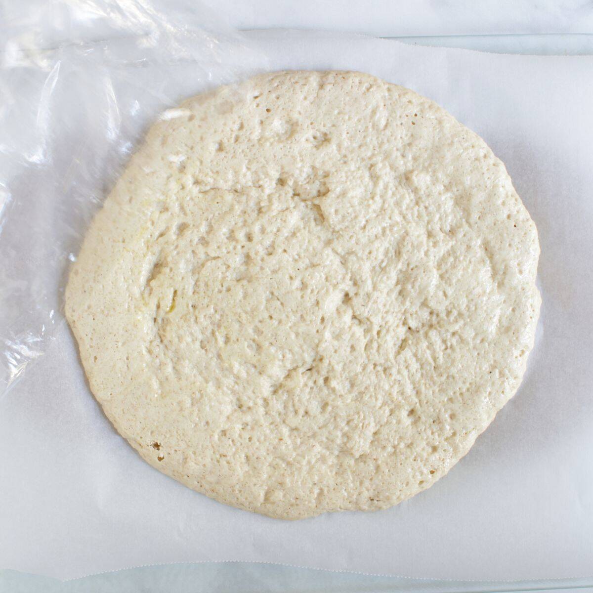 plastic wrap being peeled from risen gluten-free pizza dough.