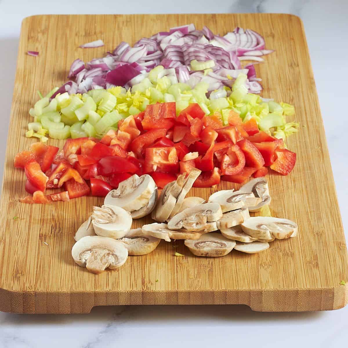 chopped mushrooms, red bell pepper, celery, and red onion on a wooden cutting board.