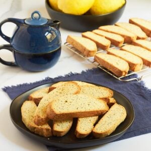 gluten-free poppyseed biscotti on black plate with blue teapot in background.