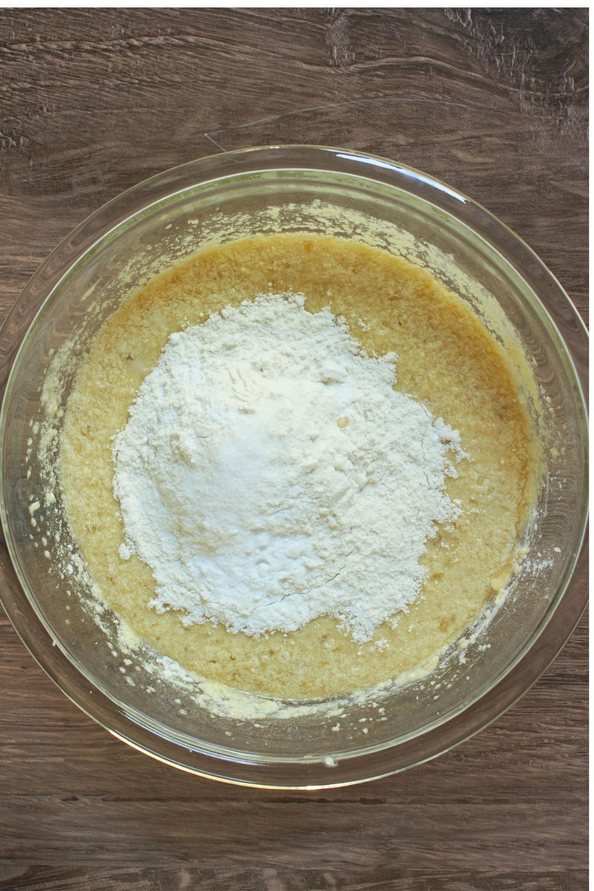 white flour piled on top of yellow, liquid batter in glass bowl.